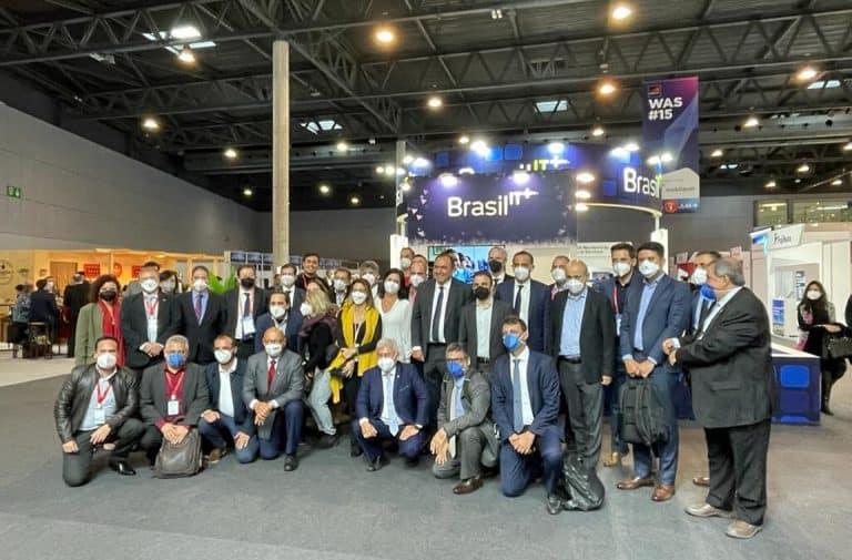 Foto de um grupo de pessoas em evento de negócios com expositores e palco ao fundo, todos de terno e máscara facial.